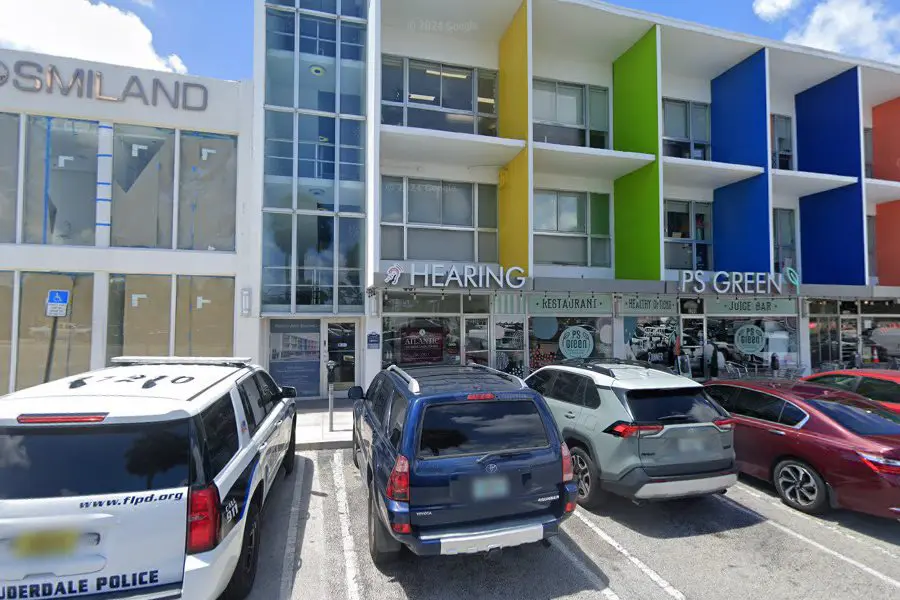 Three-story building with colorful vertical panels, restaurants and shops on the ground floor, and parked cars in front. Visible signs include HEARING and PS GREEN. The sky is partly cloudy.