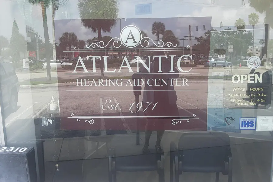 A glass storefront sign reads “Atlantic Hearing Aid Center, Est. 1971.” The reflection shows palm trees, cars, and a person taking the photo. A door sign lists open hours: Mon–Thurs 8–4, Fri 8–2.