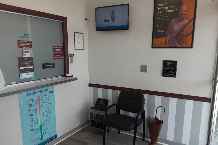 Small waiting area with a black chair, umbrella, phone, and wall-mounted TV. A window with signs is on the left, a daily noise chart below, and a tennis-themed poster on the wall. The floor is wood-style.