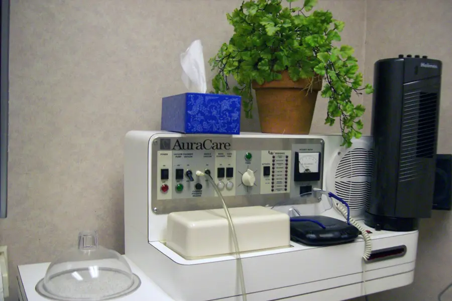 A medical device labeled AuraCare sits on a counter, surrounded by a tissue box, a potted plant, and other equipment in a clinical room. A plastic dome and some cables are also visible.