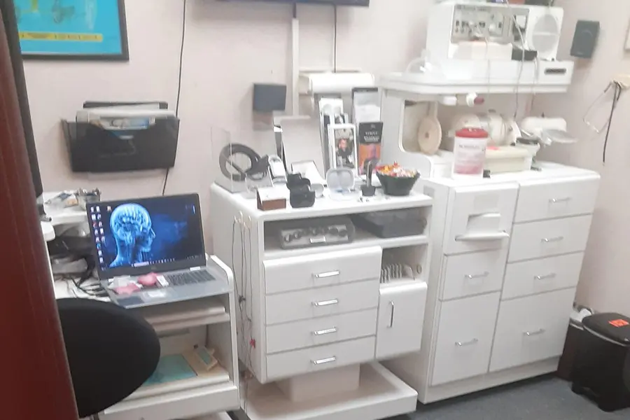 A medical office with white cabinets, drawers, medical supplies, and equipment. A laptop displaying a brain scan is on a small desk with a chair. There are posters and various items organized throughout the room.