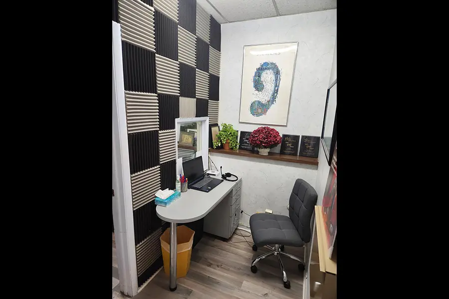 A small modern office with a curved desk, laptop, and chair. The wall is decorated with acoustic foam panels, books, red flowers, and a framed artwork featuring a large blue question mark.