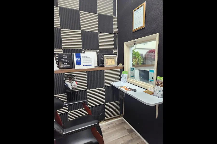 A small room with black and white soundproof foam panels, a black chair, a shelf with brochures and headphones, and a window with a counter looking into another area.