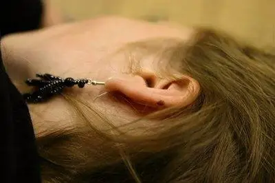 A close-up of a persons ear with a dangling black beaded earring and a small mole visible on the ear, while they are lying on their side with brown hair partially covering their face.