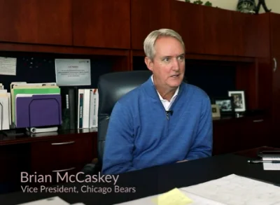 A man in a blue sweater sits at an office desk with paperwork and folders. Text on the image reads, Brian McCaskey, Vice President, Chicago Bears.