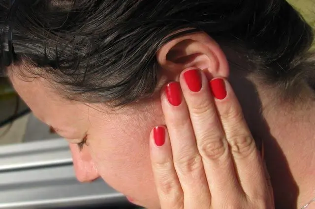A person with short, dark hair touches their ear with their hand, showing red-painted nails. The person’s eyes are closed, and sunlight highlights their face and hand.